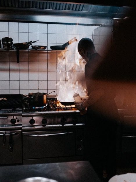 A chef cooking in a commercial kitchen with flames over the frying pan represents the WELL air concept