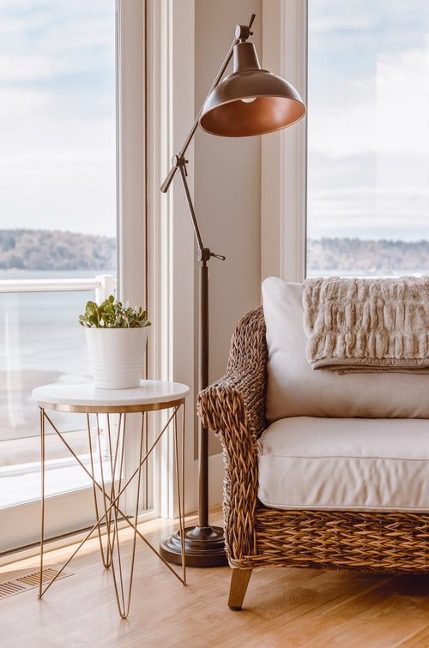 A copper floor lamp beside a woven wicker chair and brass side table overlooking a waterfront vista represents the thermal comfort concept